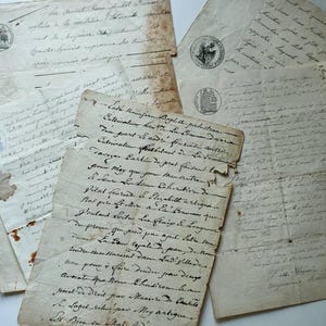 May include: A collection of antique, off-white paper documents featuring handwritten cursive French text in dark ink. The papers show signs of age with frayed edges, brown staining, and several circular ink stamps with detailed illustrations.