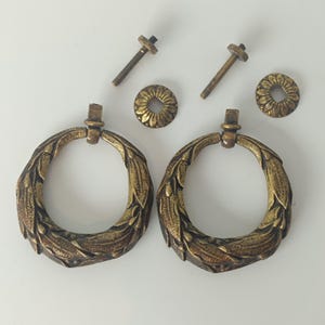 Op de afbeelding: Twee ringgrepen van antiek messingkleurig metaal met een gedetailleerd bladpatroon. De set bevat twee bijpassende ronde achterplaten met een zonnestraalontwerp en twee metalen bouten met schroefdraad voor installatie op een vlak oppervlak.