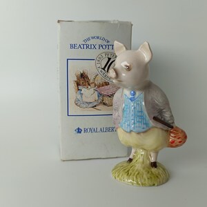 Peut inclure: Une figurine en porcelaine représentant un cochon vêtu d'un gilet à carreaux bleus, d'un pantalon jaune et d'une veste grise, posée sur un socle vert. À côté se trouve une boîte rectangulaire blanche marquée The World of Beatrix Potter et Royal Albert.