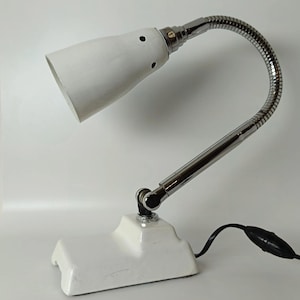 Könnte beinhalten: Eine weiße Schreibtischlampe mit einem glänzend weißen Sockel und einem passenden weißen konischen Lampenschirm mit kleinen Belüftungslöchern. Die Lampe hat einen flexiblen, verchromten Schwanenhalsarm und eine gerade Chromstange.