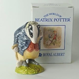 Peut inclure: Une figurine en céramique représentant un blaireau portant une veste bleue, un gilet rouge et un pantalon jaune, debout sur une base verte. Derrière se trouve une boîte en carton blanche marquée The World of Beatrix Potter et Royal Albert.