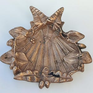 May include: A metallic bronze-toned dish shaped like a scallop shell, featuring a raised butterfly at the top center and detailed embossed leaves and small berries along the curved edges, with a textured, radiating ribbed pattern on the interior surface.