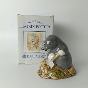 Puede incluir: Una figura de cerámica de un topo gris sentado sobre un montículo marrón texturizado con hojas verdes, acompañada de una caja de cartón blanca con la etiqueta The World of Beatrix Potter y Royal Albert con una ilustración.