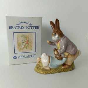 Op de afbeelding: Een keramieken beeldje van twee konijnen, één staand in een paars jasje en één liggend in een blauw hemd, op een groene basis. Daarnaast staat een witte doos met het opschrift THE WORLD OF BEATRIX POTTER en ROYAL ALBERT met een illustratie.