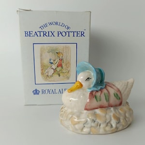 Peut inclure: Une figurine en céramique représentant un canard blanc portant un bonnet bleu et un châle rose, posé sur une base en forme de nid texturé. Derrière la figurine se trouve une boîte en carton blanche marquée The World of Beatrix Potter.