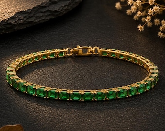 Könnte beinhalten: Ein goldfarbenes Tennisarmband mit einer durchgehenden Reihe quadratisch geschliffener, leuchtend grüner Edelsteine. Das Armband wird mit einem Metall-Kastenschloss gesichert und zeigt ein einheitliches Design aus Gliedern.