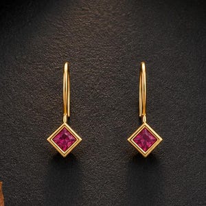 May include: A pair of gold drop earrings featuring a square-cut, vibrant pink gemstone set in a simple gold frame. The earrings hang from a thin, curved gold wire hook, showcasing a clean, geometric design that highlights the deep pink color.