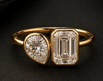 Könnte beinhalten: Ein Goldring mit zwei verschiedenen Steinen in Zargenfassung. Ein Stein ist im Birnenschliff gehalten, der andere im rechteckigen Smaragdschliff. Die Ringschiene besteht aus glattem, poliertem Goldmetall, das beide Steinfassungen verbindet.