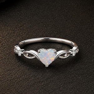 Può includere: Un anello color argento con una pietra preziosa opale a forma di cuore con finitura iridescente. La fascia presenta un delicato design in metallo intrecciato su entrambi i lati del cuore, accentuato da piccole pietre chiare incastonate.