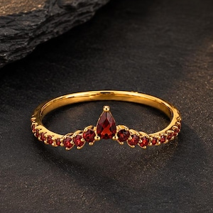 May include: A gold ring featuring a V-shaped band set with multiple small round red gemstones and a single central pear-shaped red gemstone. The band has a polished metallic finish and follows a chevron silhouette with stones along the front.