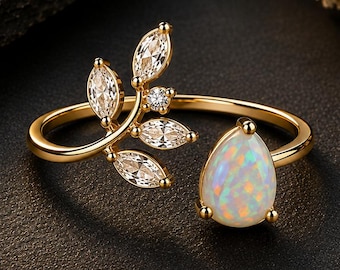 May include: A gold open ring featuring a pear-shaped iridescent opal on one end and a branch-like design on the other. The branch is adorned with five marquise-cut clear stones and one small round clear stone, creating a delicate botanical aesthetic.