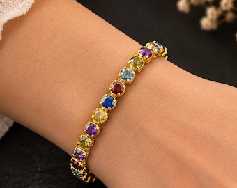 Könnte beinhalten: Ein goldfarbenes Tennisarmband mit einer Reihe von rund geschliffenen, mehrfarbigen Edelsteinen in Blau, Rot, Gelb, Grün und Violett. Jeder Stein ist in einer Krappenfassung gefasst, was eine durchgehende Linie aus lebendigen Farben erzeugt.