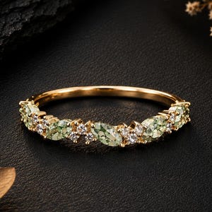 May include: A gold band ring featuring a series of alternating marquise-cut light green gemstones and small round clear stones set along the top half of the band. The metal has a polished finish, and the stones are arranged in a continuous row.