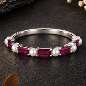 Peut inclure: Une bague argentée présentant un motif alterné de pierres rectangulaires rouge foncé taillées en baguette et de petites perles blanches rondes serties le long de l'anneau. L'anneau en métal est poli et lisse, avec une disposition symétrique.