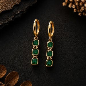 May include: A pair of gold hoop earrings, each featuring a vertical drop component with three square-cut, deep green gemstones. The stones are set in gold frames and connected by small gold beads, creating a linear, dangling design.
