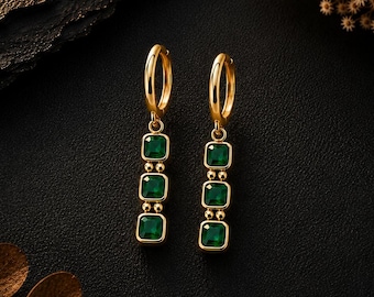 May include: A pair of gold hoop earrings, each featuring a vertical drop component with three square-cut, deep green gemstones. The stones are set in gold frames and connected by small gold beads, creating a linear, dangling design.