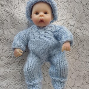 May include: A small doll wearing a light blue knitted outfit, including a hat and a one-piece suit. The doll has a realistic face with brown eyes and is posed on a white lace surface. The knitted clothing appears soft and handmade.