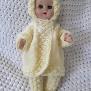 May include: A vintage baby doll wearing a pale yellow knitted outfit. The outfit includes a hooded jacket with three decorative buttons and matching pants. The doll is posed on a white, textured blanket.