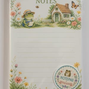 May include: A rectangular notepad with horizontal lines and the word NOTES at the top. The design features a green frog in a straw hat, a small cottage, flowers, a bee, and a butterfly. A circular sticker shows a frog and the text NOTEPAD.