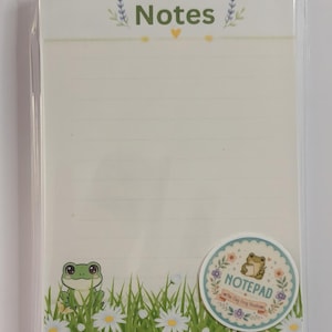 May include: A rectangular notepad with cream-colored lined paper. The top features the word Notes in green with lavender sprigs. The bottom displays a green grassy field with white daisies and a green frog. A circular sticker reads NOTEPAD.