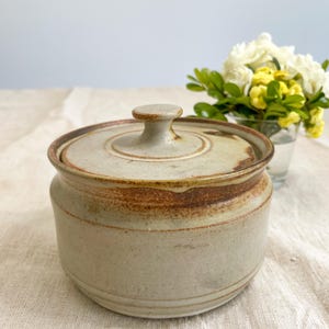 May include: A cylindrical ceramic jar with a matching lid, featuring a speckled light grey glaze and a prominent band of earthy brown, rust-coloured glaze around the upper rim and the lid edge, with a small knob handle centred on the lid.