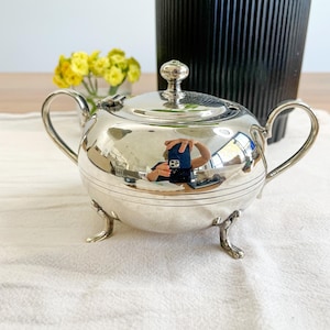 May include: A polished silver-toned sugar bowl with a rounded body, a matching lid with a small knob handle, two curved side handles, and three decorative feet. The surface is reflective, showing a distorted view of the surrounding environment.