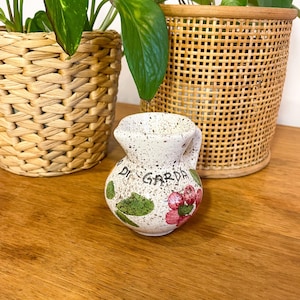 May include: A small, speckled ceramic pitcher with a rounded body and a single handle. The off-white surface features a hand-painted red flower with green leaves and the black text "DI GARDA" written across the front of the vessel.