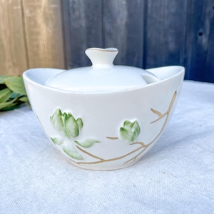 May include: A white ceramic sugar bowl with a matching lid, featuring a raised relief design of green magnolia flowers and gold-painted branches. The bowl has a curved, wide-rimmed shape with a small notch on the side for a spoon.