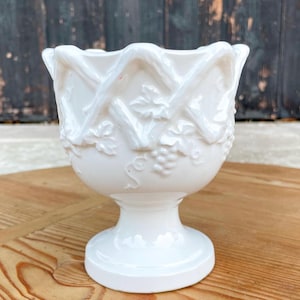 May include: A white ceramic pedestal bowl featuring a raised relief pattern of intertwined vines and grape clusters around the exterior. The bowl has a scalloped rim and a circular base, with a glossy finish that reflects light across the surface.