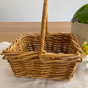 May include: A rectangular woven wicker basket featuring a light brown natural finish and a single arched handle. The basket is constructed with a sturdy base and woven sides, showcasing the organic texture and pattern of the willow material.