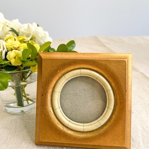 May include: A square gold-toned picture frame with a circular centre opening. The circular inner border is off-white with thin, dark, evenly spaced vertical lines. The frame has a smooth, matte finish and a slightly weathered, textured appearance.