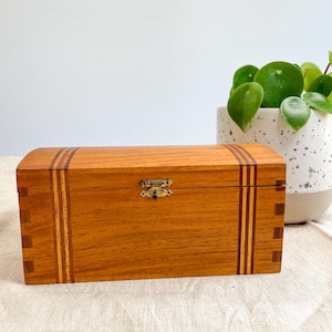 May include: A rectangular wooden box with a curved lid, featuring a light brown wood grain, darker vertical wood inlay stripes, and a small brass-coloured metal latch centred on the front. The box has visible finger joints at the corners.