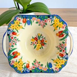 May include: A square ceramic bowl with two side handles, featuring a cream-coloured interior and a vibrant floral pattern. The design includes yellow daffodils, red and blue tulips, and pink blossoms, all bordered by a bold blue rim and green leaves.