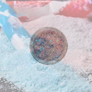 May include: A small, round, clear container filled with a mixture of fine, multi-colored glitter particles. The blend includes shades of pink, gold, and iridescent blue, with larger hexagonal blue sequins scattered throughout the textured contents.