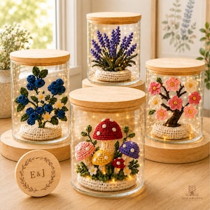 Peut inclure: Quatre bocaux en verre avec couvercles en bois contenant des plantes crochetées : un myrtillier, un brin de lavande, un cerisier en fleurs et un groupe de champignons colorés. Un couvercle en bois séparé porte une couronne et les lettres E&J.
