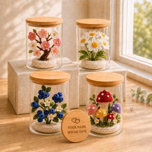 Peut inclure: Quatre bocaux en verre transparent avec couvercles en bois, contenant chacun une plante crochetée : un cerisier rose, des marguerites blanches, des myrtilles bleues et des champignons colorés. Un disque en bois gravé de deux cœurs et du texte YOUR NAME SPECIAL DATE.
