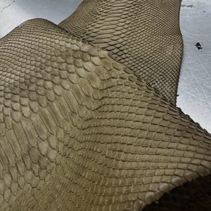 Pode incluir: Um pedaço de couro de pele de cobra na cor castanho-claro com um padrão de escamas distinto e repetitivo na superfície. O material está dobrado, mostrando textura e profundidade. O texto 'REPTILE SKIN TANNERY' é visível no canto superior esquerdo.