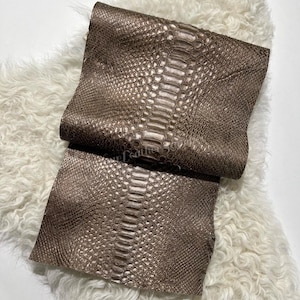 May include: A rectangular piece of metallic bronze-colored leather featuring a prominent, raised scale pattern that mimics the texture of a snake. The material has a distinct central spine of larger, elongated scales flanked by smaller, rounded scales.