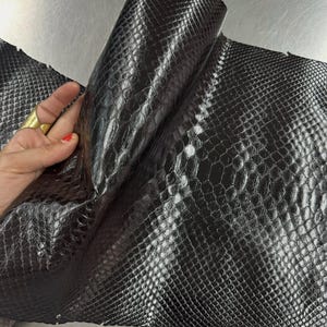Peut inclure: Une feuille de cuir de serpent noir avec une finition brillante et un motif d'écailles répétitif distinct. Le matériau est flexible et replié pour révéler la texture et l'éclat de la peau. Le texte Reptile Skin Tannery est visible.
