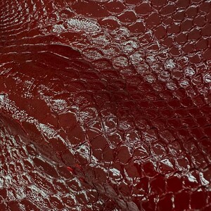 May include: A deep red, glossy material featuring a textured, raised scale pattern resembling reptile skin. The top left corner displays the white text 'REPTILE SKIN TANNERY' with an ornate decorative flourish above the brand name.