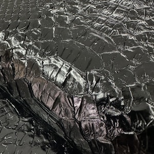 May include: A glossy black reptile skin hide featuring a distinct, raised scale pattern with a reflective finish. The surface displays a natural, textured grid of interlocking scales. The text "REPTILE SKIN TANNERY" is visible in the top left corner.