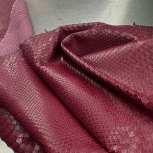 Puede incluir: Una piel de serpiente de color burdeos intenso con una textura de escamas en relieve. El cuero está doblado, mostrando el patrón natural y el rico color rojo oscuro. Un logotipo en la esquina superior izquierda dice Reptile Skin Tannery.
