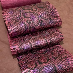 Può includere: Un rotolo di pelle con motivo a pelle di serpente rosa e nero con una superficie testurizzata e squamata. Il materiale presenta un motivo ripetitivo a rombi nelle tonalità magenta e viola intenso su una base di pelle color malva opaco.