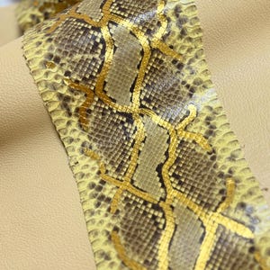 May include: A strip of textured snakeskin leather featuring a natural brown and yellow scale pattern overlaid with metallic gold foil accents, layered over a smooth, solid light beige leather surface.