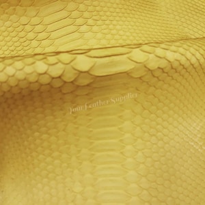 May include: A bright yellow leather sheet featuring a distinct, raised snakeskin texture with a repeating scale pattern. The surface displays a semi-gloss finish and includes the watermark text Your Leather Supplier across the centre.