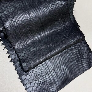 May include: A piece of black snakeskin leather with a distinct, repeating scale pattern. The surface has a subtle sheen and a textured, reptilian appearance. The text Your Leather Supplier is printed in a light color across the center.