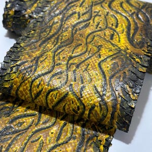 May include: A rolled piece of textured material featuring a vibrant yellow and black snake-skin pattern. The surface displays a repeating, organic wavy design with deep black grooves contrasting against the bright yellow scales.