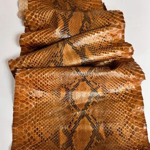 May include: A long, rectangular piece of snakeskin leather featuring a natural tan and brown diamond-shaped scale pattern. The material has a glossy, textured surface with overlapping scales that create a distinct, repeating geometric design.