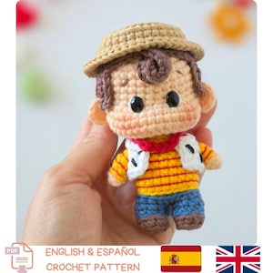 May include: A small crocheted doll featuring a tan straw-style hat, brown hair, and black bead eyes. The figure wears a yellow and orange striped shirt, a white vest with black accents, a red neckerchief, blue pants, and brown shoes.