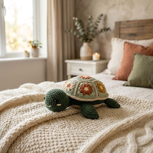 May include: A crochet turtle toy featuring a dark green head and limbs with a shell constructed from light green hexagonal patches, each centred with a small flower motif in shades of white, orange, and yellow, resting on a textured cream blanket.